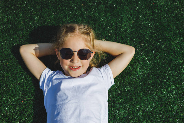 8 yaşında, güneş gözlüklü neşeli bir kız parkta çimlerin üzerinde yatıyor. Çocuk dışarıda. yaz parkında dinlen. Yukarıdan bak. Bir yazıt için yer - Fotoğraf, Görsel