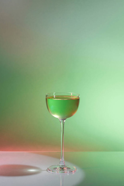 Gin koktél Mátrix. Egy zöld színű gin-alapú koktél, amit egy magas üvegszárú vermut pohárba öntöttek. Az üveg mellett egy átlátszó folyadékkal és zöld festékkel ellátott főzőpohár oldódik. Egy üveg és egy főzőpohár áll egy üveg háttér. - Fotó, kép