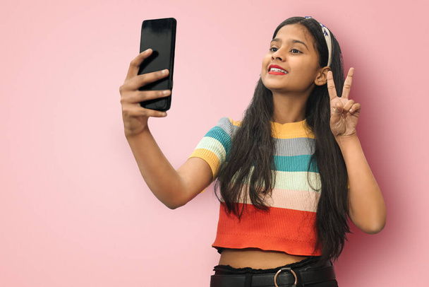 Portrait of pleased nice lovely attractive smiling indian asian girl posing isolated wearing t-shirt doing selfie shot on mobile cell phone  with victory or peace gesture. Mock up copy space. - Fotó, kép