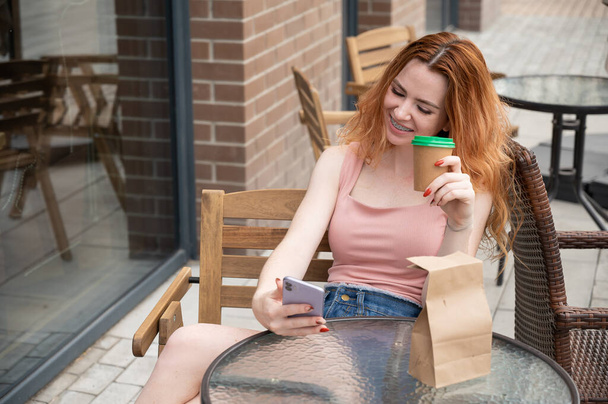 Young red-haired woman with braces on her teeth drinks coffee on a summer veranda - Foto, imagen