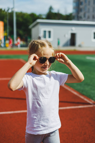 Egy szőke kislány fut a sportpályán és mosolyog, napszemüveget és fehér pólót visel. Nyaralás és nyaralás. - Fotó, kép