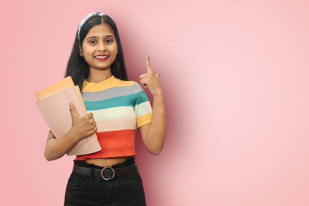 Excited young indian asian student girl posing islolated holding textbooks and pointing up with back right hand, looking directly at camera, dark haired female expressing positive emotions. - Photo, Image