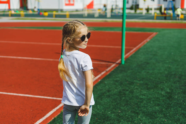 Маленька блондинка біжить на спортивному полі і посміхається, вона носить сонцезахисні окуляри і білу футболку. Літні канікули та відпустка
. - Фото, зображення