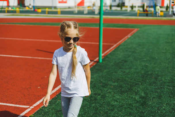 Маленька блондинка біжить на спортивному полі і посміхається, вона носить сонцезахисні окуляри і білу футболку. Літні канікули та відпустка
. - Фото, зображення