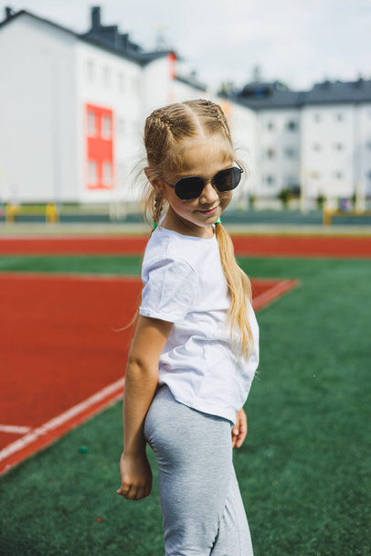 A little blonde girl is running on the sports field and smiling, she is wearing sunglasses and a white T-shirt. Summer holidays and vacation time. - Foto, afbeelding