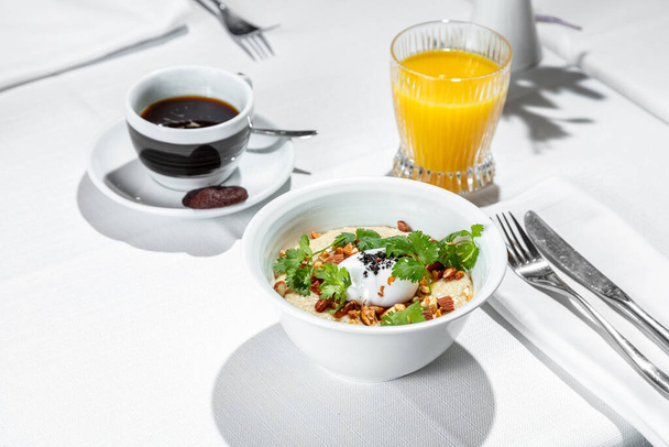 Breakfast. Morning dish of salted oatmeal with almonds, poached eggs and parsley leaves. Porridge with ingredients lies in a white, ceramic, deep bowl. The bowl is on a table with a white tablecloth. Next to the porridge is a glass of orange juice an - Foto, Imagen