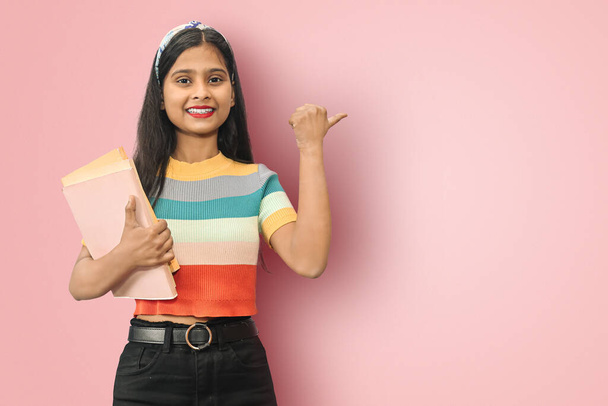 Excited young indian asian girl student posing islolated holding textbooks and pointing thumb up to copy space, looking directly at camera, dark haired female expressing positive emotions. - Photo, Image
