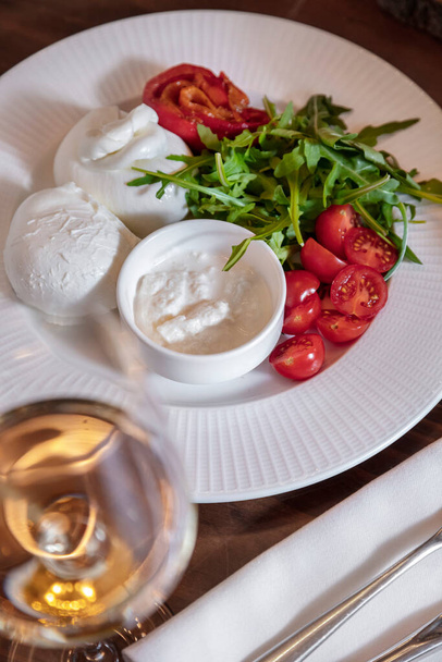 Salad with tomatoes, meat, mozzarella, arugula, sour cream and sauce in a plate on a wooden table with a glass of wine and cutlery - Photo, image