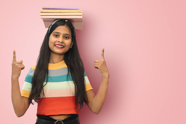 Excited young indian asian girl student posing islolated balancing books on her head and  pointing up with both hands to a copy space, looking directly at camera expressing positive emotions - 写真・画像