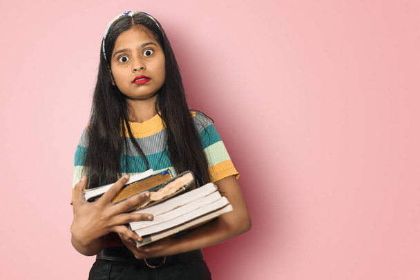 Portrait of a amazed indian asian girl student with open mouth and big eyes posing isolated holding a lot of books and shocked by seeing exam syllabus. Education concept - 写真・画像