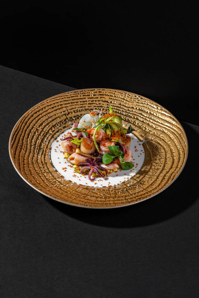 Warm salad with seafood. Salad of octopus, squid, shrimp, scallop, sesame seeds in paprika, lettuce and tartar sauce on Japanese mayoness. Salad lies on a golden plate with wide margins. The plate stands on a black paper background. - Фото, изображение
