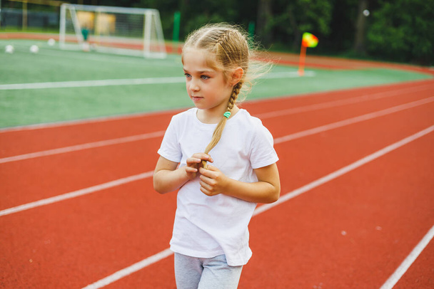 A little girl of 7-8 years old in a white t-shirt runs outdoors in the stadium. The girl is doing sports on a sunny day - 写真・画像