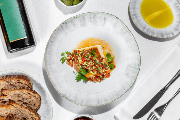 Italian dish. Sea bass fillet fried in a grill pan with vegetable tabouleh. The food lies in a light handmade ceramic plate. Nearby is a bowl of olives, a saucer of olive oil, a plate of buckwheat bread and grissini, and a dark bottle of olive oil wi - Photo, Image