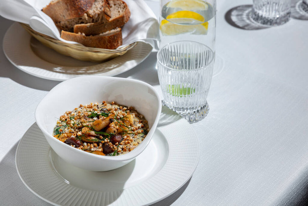 Gachas con trigo, champiñones y hierbas en un plato sobre la mesa con un vaso de agua y un plato de pan - Foto, Imagen