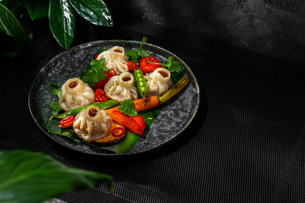 Japanese Jiaozi dumplings with cheese. Dumplings, rounded, boiled and fried in a wok. Nearby lies green peas, microgreen sprouts, chili peppers and carrots. The food lies in a round, gray, ceramic plate. The plate stands on a wicker, black bedding. B - Photo, image