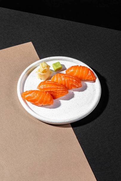 Nigiri with salmon. Large pieces of salmon sashimi lie on top of the rice. Nearby is pickled ginger and a cube of wasabi. The food lies on a round, light, ceramic plate with low sides. The plate stands on a black and beige paper background. - Fotoğraf, Görsel
