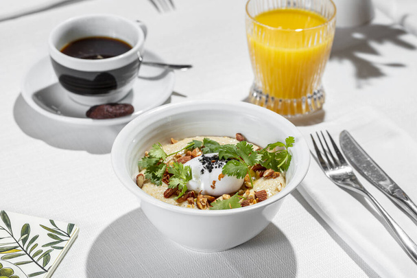 Breakfast. Morning dish of salted oatmeal with almonds, poached eggs and parsley leaves. Porridge with ingredients lies in a white, ceramic, deep bowl. The bowl is on a table with a white tablecloth. Next to the porridge is a glass of orange juice an - Fotoğraf, Görsel