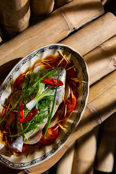 Salade met gebakken vis, chili pepers, tomaten, saus, uien, dille, en strookjes zoete aardappelen in een bord op tafel gemaakt van bamboe stammen - Foto, afbeelding