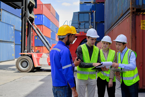 Skupina multiraciálních pracovníků v bezpečnostních uniformách a s pevnými klobouky pracuje v logistickém terminálu s mnoha stohy kontejnerů, kontrola nakládky zboží pro nákladní dopravu. - Fotografie, Obrázek