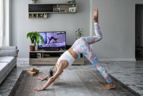 junge Frau macht Online-Yoga-Übungen zu Hause und schaut Lehrer-Tutorial auf dem Fernsehbildschirm an - Foto, Bild