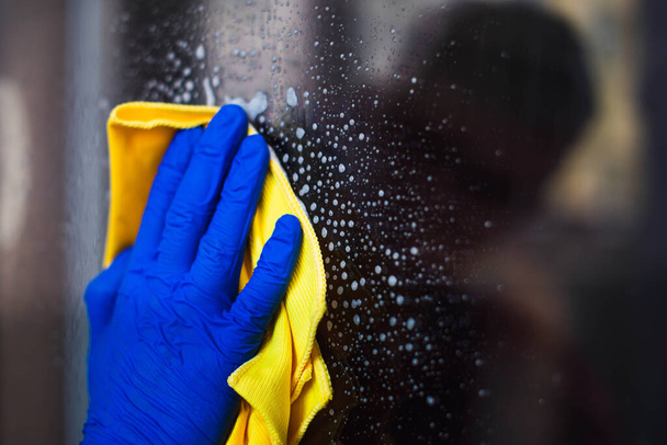 A woman or man washes a window at home. House cleaning. Washing dirty window glass detergent for window washing wearing blue mittens and a yellow cloth rag - Valokuva, kuva