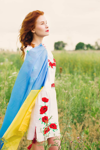 A beautiful Ukrainian woman in a folk dress with a flag of Ukraine is walking along the field - Foto, imagen