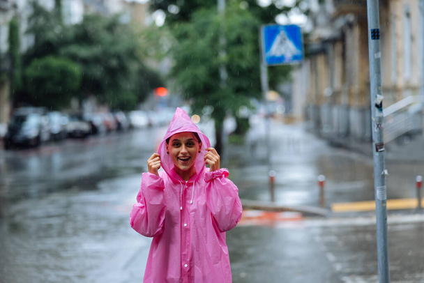 Junge lächelnde Frau mit rosa Regenmantel auf der Straße, während sie an einem regnerischen Tag einen Spaziergang durch die Stadt genießt. - Foto, Bild