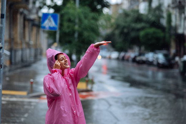Jonge lachende vrouw met een roze regenjas op straat terwijl ze geniet van een wandeling door de stad op een regenachtige dag. - Foto, afbeelding