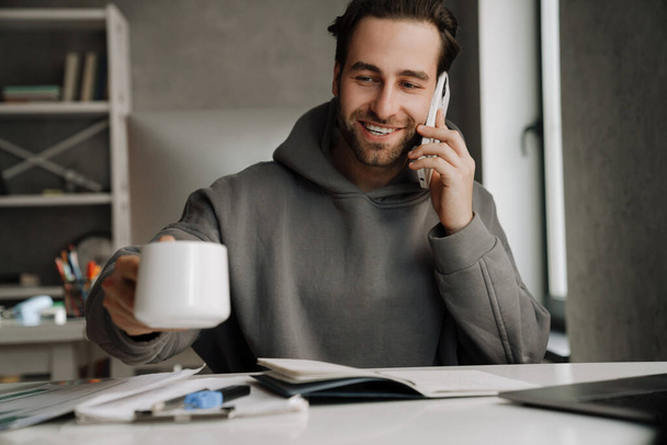 Young beard man talking on cellphone while drinking coffee in office - Photo, Image