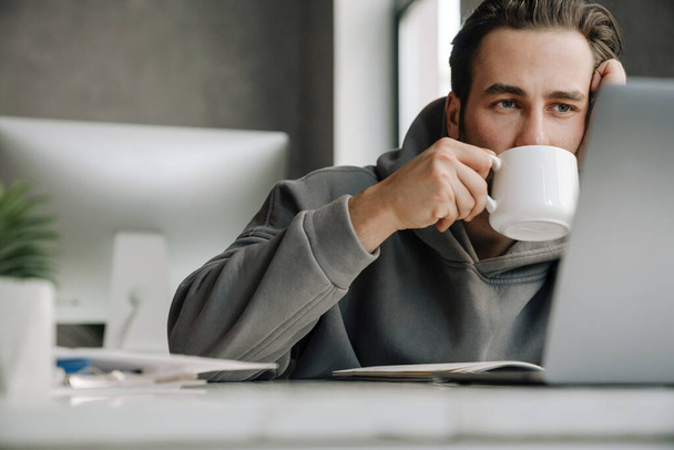 Young beard man drinking coffee while working with laptop in office - Photo, image