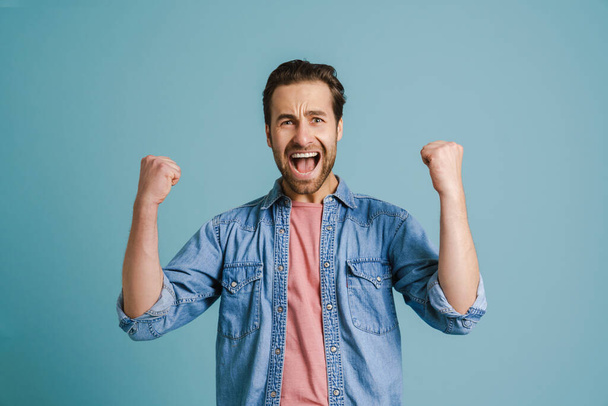 Young excited man screaming while making winner gesture isolated over blue background - Foto, Imagem