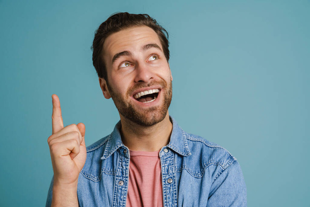Young excited man wearing shirt smiling and pointing finger upward isolated over blue background - Foto, Imagen