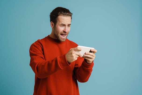 Young excited man screaming while playing online game on cellphone isolated over blue background - Fotografie, Obrázek