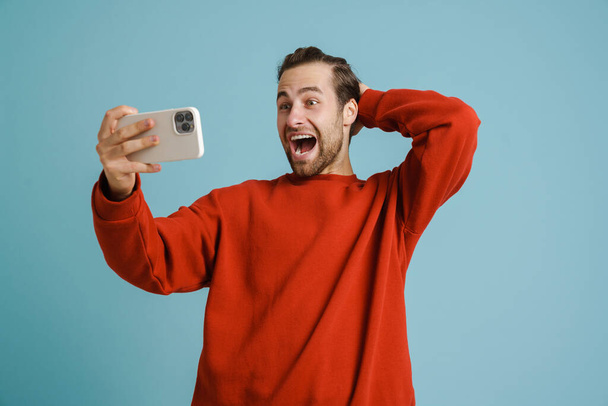 Young excited man screaming and taking selfie on cellphone isolated over blue background - Foto, immagini