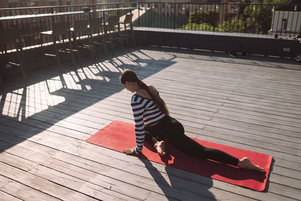 Young sporty happy woman in sportswear doing stretching exercises on yoga mat on house roof in early morning - Photo, image