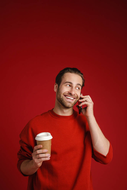 Young bristle man drinking coffee while talking on cellphone isolated over red background - 写真・画像