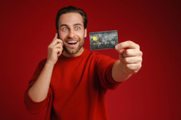 Surprised man showing credit card while talking on cellphone isolated over red background - Foto, Imagen