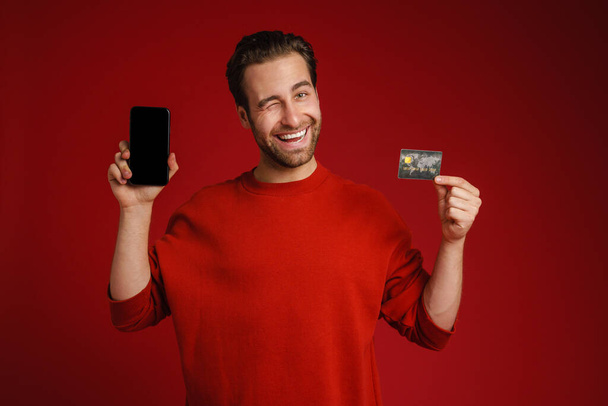 Überraschter Mann zwinkert und zeigt Kreditkarte und Handy isoliert über rotem Hintergrund - Foto, Bild