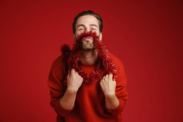 Молодий чоловік з щетини сміється, розважаючись з мішурою ізольовано на червоному тлі
 - Фото, зображення