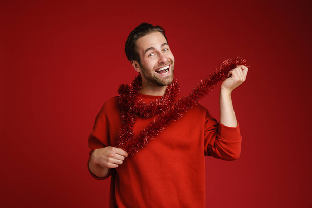 Young bristle man laughing making fun with tinsel isolated over red background - Fotografie, Obrázek
