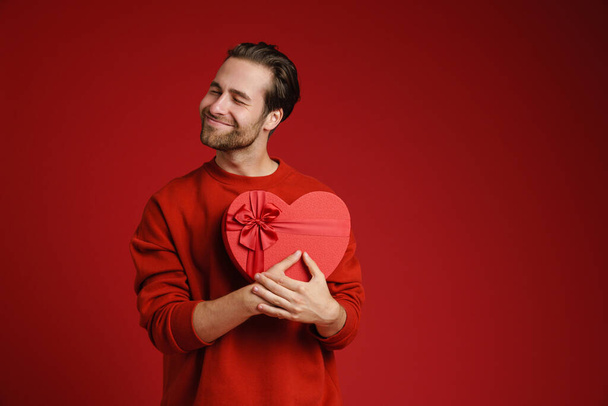 Young bristle man smiling while posing with heart gift box isolated over red background - Φωτογραφία, εικόνα