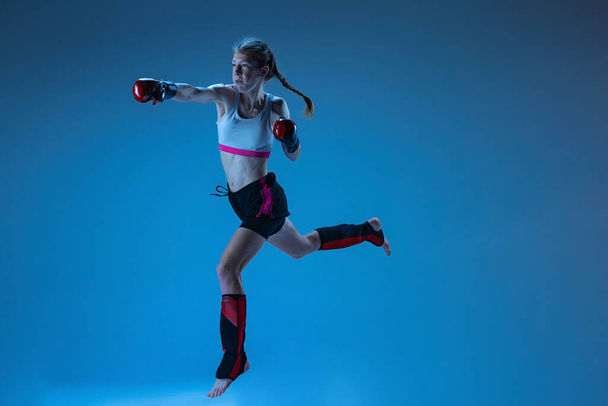 Fight. Dynamic portrait of young girl, junior MMA fighter in action, motion isolated on blue background in neon light. Concept of sport, competition, action, achievements. Copy space for ad. - Фото, зображення