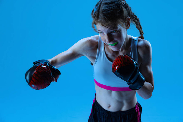 Attack. Dynamic portrait of young girl, junior MMA fighter in action, motion isolated on blue background in neon light. Concept of sport, competition, action, achievements. Copy space for ad. - Photo, Image