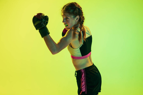 Dynamic portrait of athletic girl, MMA fighter training isolated on gradient yellow-green background in neon. Concept of sport, competition, action, achievements. - Photo, image