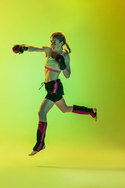 Punch in jump. Dynamic portrait of athletic girl, MMA fighter training isolated on gradient yellow-green background in neon. Concept of sport, competition, action, achievements. - Foto, imagen