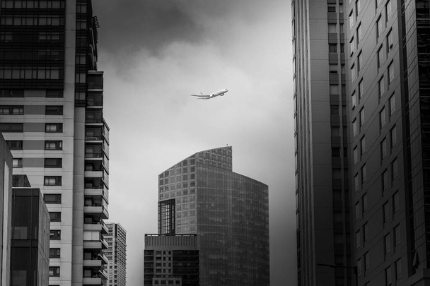 A grayscale of a plane flying high in the sky between modern skyscrapers - Foto, immagini