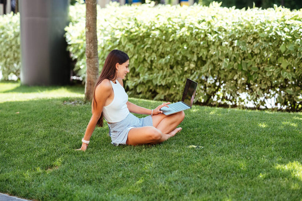 The concept of remote work. A young girl works with a laptop in the fresh air in the park, sitting on the lawn. - 写真・画像