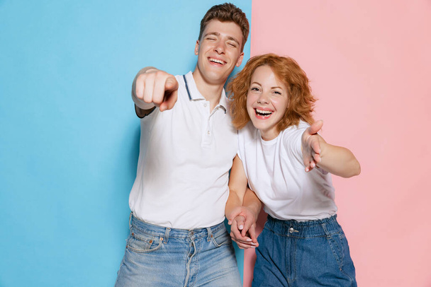 Portrait of young couple, posing, laughing and pointing with fingers at camera isolated over pink blue studio background. Concept of youth, emotions, facial expression, love, relationship. Poster, ad - Photo, image