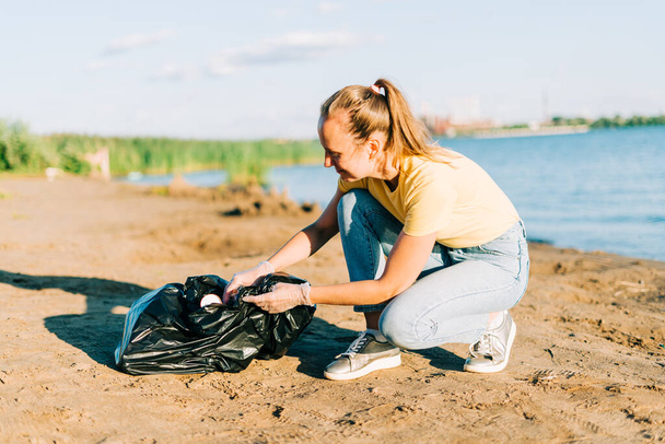 Young female volunteer satisfied with picking up trash, a plastic bottles and coffee cups, clean up beach with a sea. Woman collecting garbage. Environmental ecology pollution concept. Earth Day. - Фото, изображение