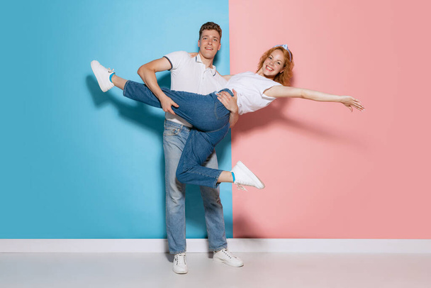 Portrait of young couple, man circling woman in his hands, posing isolated over pink blue studio background. Concept of youth, emotions, facial expression, love, relationship. Poster, ad - Foto, Bild